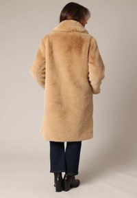 Abrigo de pelaje sintético beige con un cuello alto, que presenta una textura suave y mangas oversize, combinado con jeans oscuros y botas de tacón negras.