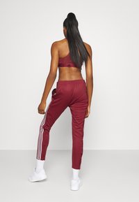 Bordeaux roter Sport-BH und passende Trainingshose mit weißen Seitenstreifen. Glattes Material, taillierte Hose, elastischer Bund und Druckknopfdetails. Weiße Sneakers.