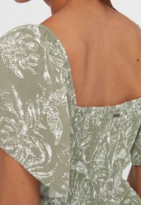 Vestido verde con estampado, con textura fruncida, que presenta mangas anchas y un respaldo cuadrado. La tela tiene un estampado floral en blanco.