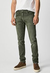 Olive green jeans made of denim, slim fit with front pockets and a single back pocket. Paired with brown suede sneakers.