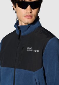 Fleecejacke mit einem blauen unteren Abschnitt und einem schwarzen oberen Abschnitt. Verfügt über eine mit Reißverschluss versehene Tasche und weiße Textelemente auf der Brust.