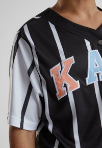 Juodai balta raištelių maikutė su V formos iškirpte, papuošta spalvotais siuvinėjimais "K", "A" ir "K". Pagaminta iš tekstūruoto audinio.