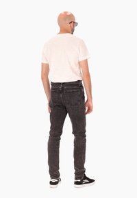 Un homme portant un jean noir, un t-shirt blanc, des baskets noires et des lunettes de soleil se tient de dos devant un fond blanc.