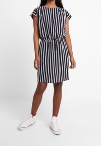 Vestido a rayas azul marino y blanco con mangas cortas, cintura fruncida y detalle de corbata. Combinado con zapatillas blancas para un look casual.
