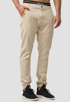Pantalones jogger de algodón beige con un diseño ajustado, un bolsillo lateral y una cinturilla negra en contraste. Llevados con zapatillas negras.