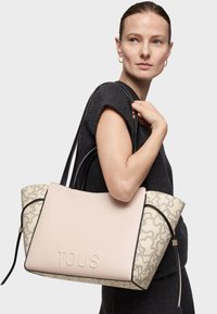 Bolso de mano en tonos beige y crema con tejido texturizado, ribete negro y logo en relieve "TOUS". Presenta asas dobles y un patrón intrincado.