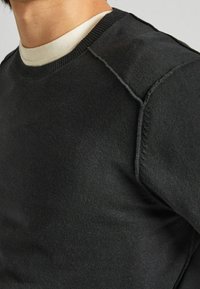 Närbild av en person som bär en svart tröja med rund hals över en vit t-shirt med rund hals, som visar detaljer kring axlar och halsringning.