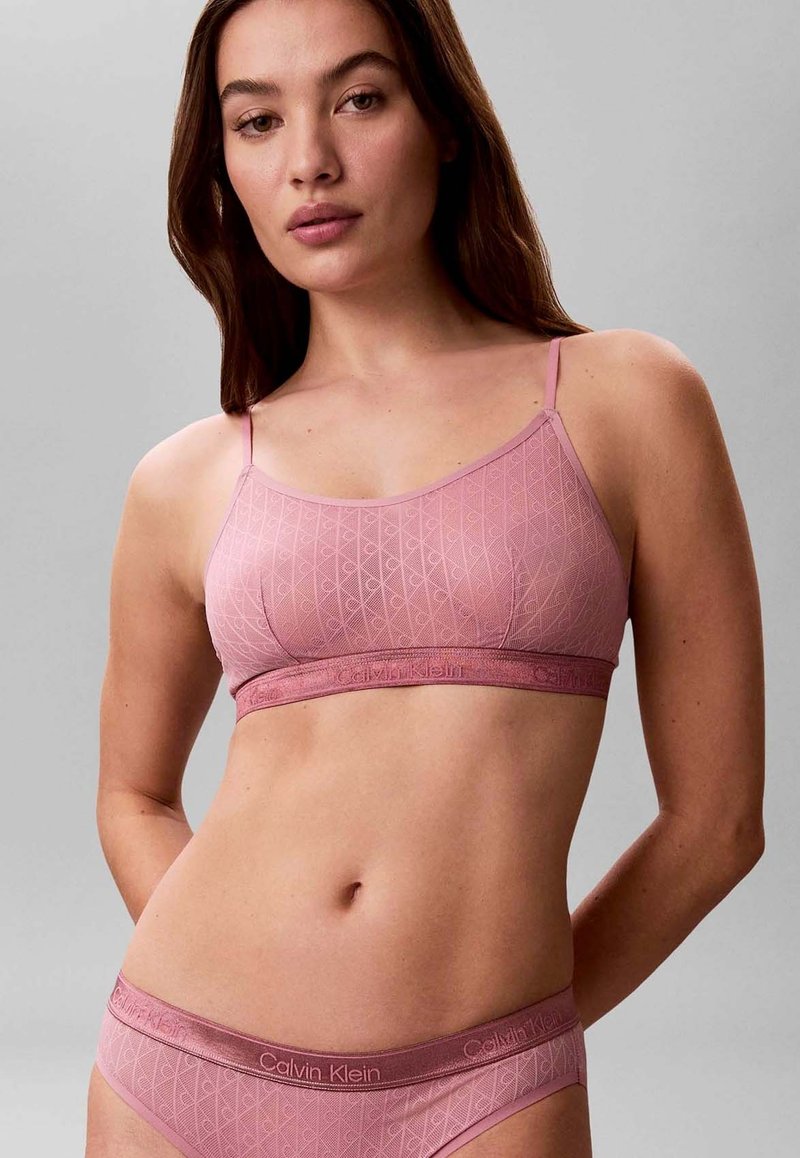 Femme portant un ensemble assorti de bralette et sous-vêtements Calvin Klein roses avec un motif géométrique subtil, posant les bras derrière le dos.