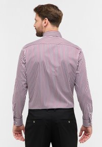 Camicia da uomo a maniche lunghe con righe verticali bordeaux e bianche, colletto classico, polsini con bottoni e pantaloni neri con due tasche posteriori.