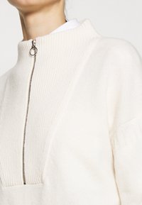 Pull en tricot crème avec col montant côtelé et fermeture éclair argentée. Présente un design de couture diagonal et des accents texturés sur les manches.