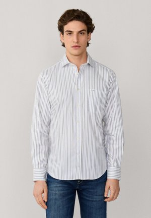 Joven vestido con una camisa blanca de botón a rayas, jeans azules y zapatos beige sin cordones, de pie frente a un fondo claro y sencillo.