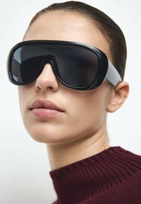 Occhiali da sole oversize neri con finitura liscia, caratterizzati da un design curvo e dettagli grigi sui lati. Indossati con un dolcevita bordeaux dalla trama lavorata.