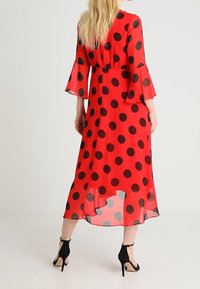 Robe rouge longueur genou en tissu transparent ornée de grands pois noirs, avec des manches évasées et une taille cintrée, associée à des talons noirs.