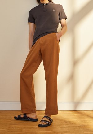 Personne portant un polo marron foncé, un pantalon large couleur rouille et des sandales noires à double bride, debout avec une main dans la poche.