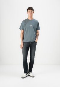T-shirt gri cu mânecă scurtă și un logo subtil pe piept, asortat cu blugi din denim închis și adidași albi cu accente gri.