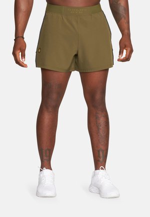 Osoba muskularna w oliwkowo-zielonych szortach Gold's Gym i białych butach sportowych, pokazująca tatuaże na nogach i przedramionach.