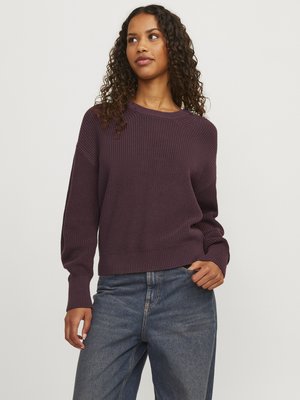 Vrouw met krullend haar, gekleed in een plomkleurige gebreide trui en blauwe jeans, staand tegen een effen lichte achtergrond.