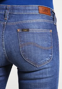 Primer plano de un bolsillo trasero de jeans Lee azules con costuras visibles y etiquetas de la marca Lee en el bolsillo y la cintura.