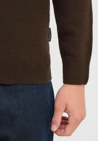 Brązowy sweter wełniany z prążkowanymi mankietami i dołem. Po boku widoczna niebieska metka z napisem "FRIDAY". Dżinsy w kolorze ciemnego denimu.