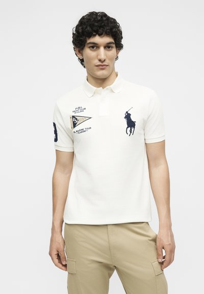 Νέος άνδρας με σκούρα σγουρά μαλλιά, ντυμένος με λευκό πόλο πουκάμισο με ναυτικό κεντημένο λογότυπο και μπεζ παντελόνια cargo, στέκεται μπροστά σε απλό φόντο.