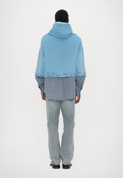 Blue oversized hooded sweatshirt with frayed hem, layered over a gray plaid shirt; paired with light wash distressed jeans and black shoes.