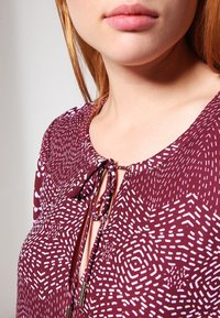 Bordeaux blouse med et geometrisk hvidt mønster, der har en V-udskæring med bindelukning og subtile metallic accenter. Glat tekstur.