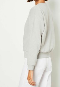 Sweat-shirt gris clair à col rond en tissu doux, avec épaules tombantes et poignets côtelés, associé à un pantalon blanc.