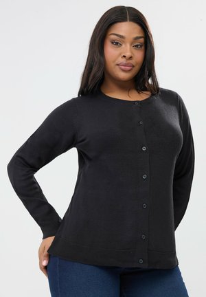 Vrouw met lang haar, gekleed in een zwart, dichtgeknoopt vest en een blauwe spijkerbroek, poserend met één hand op haar heup tegen een effen achtergrond.