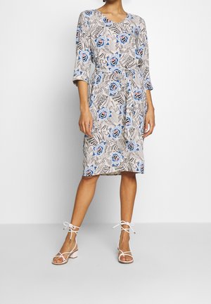 Donna con vestito beige al ginocchio con stampa floreale blu e maniche a tre quarti, abbinato a sandali bianchi con tacco a blocco e lacci.