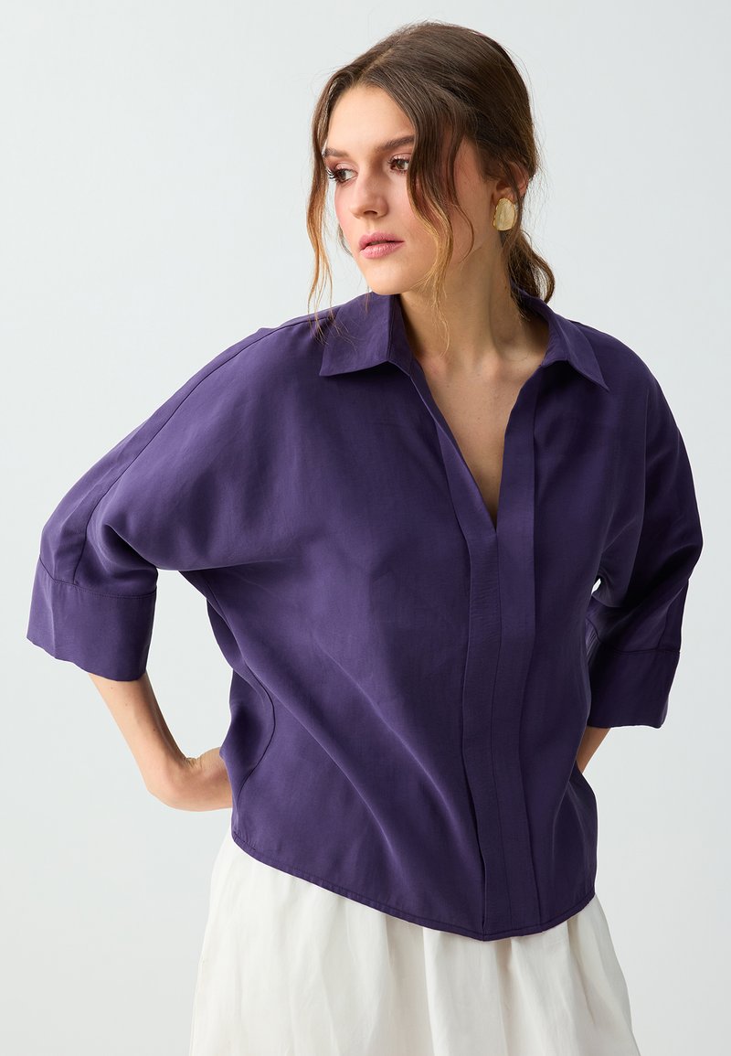 Femme aux cheveux bruns attachés porte une chemise violette ample à manches trois-quarts et une jupe blanche, regardant vers sa gauche.