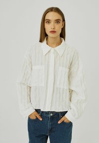 Vit, skrynklig skjorta med krage, med två framfickor och långa ärmar, i kombination med blå högmidjade jeans.