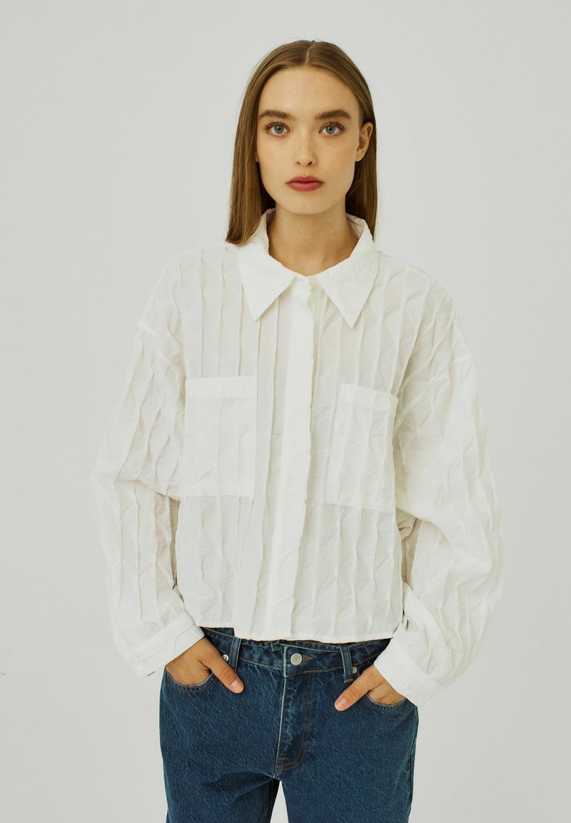 Vit, skrynklig skjorta med krage, med två framfickor och långa ärmar, i kombination med blå högmidjade jeans.