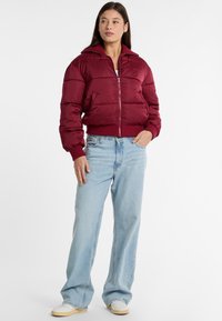 Giubbotto imbottito bordeaux con zip, design corto, orlo e polsini a coste; indossato con jeans a gamba larga blu chiaro e scarpe da ginnastica bianche.