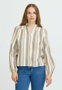 Kvinde med langt hår iført en cremefarvet bluse med multifarvede lodrette striber og blå højtaljede jeans, stående foran en ensfarvet baggrund.