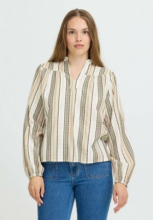 Kvinde med langt hår iført en cremefarvet bluse med multifarvede lodrette striber og blå højtaljede jeans, stående foran en ensfarvet baggrund.