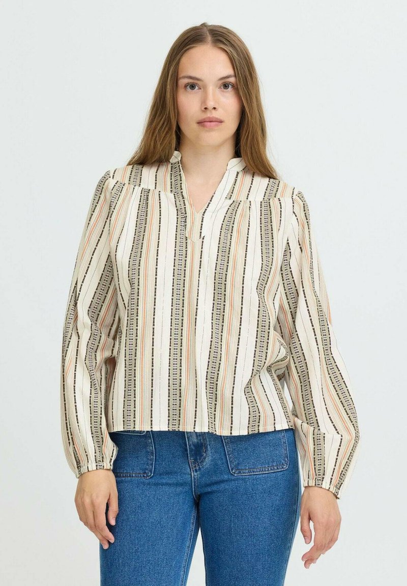 Kvinde med langt hår iført en cremefarvet bluse med multifarvede lodrette striber og blå højtaljede jeans, stående foran en ensfarvet baggrund.