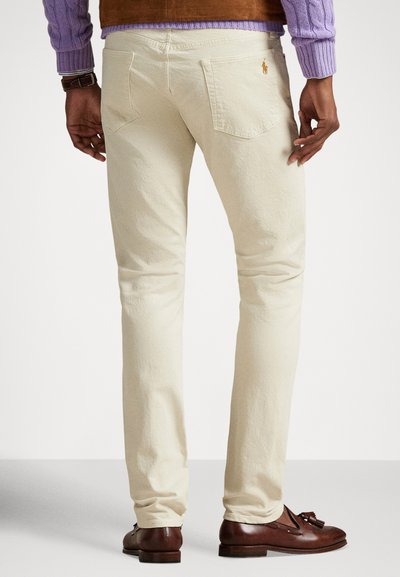 Svjetlo bež pamučne hlače uskog kroja, s stražnjim džepovima, suptilnom teksturom i malim izvezenim logotipom na leđima. Smeđe kožne cipele.