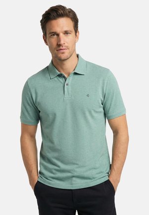 Man wearing a short-sleeve green polo shirt with collar and two buttons, standing with hands in pockets against a plain background.