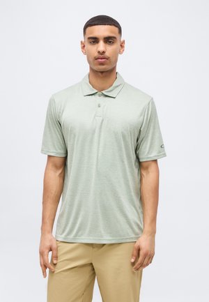 Férfi világoszöld rövid ujjú galléros pólóban és bézs nadrágban, egyszerű fehér háttér előtt, előre nézve.