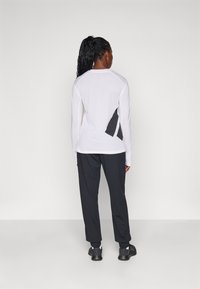Weißes langärmliges Sportoberteil aus strukturiertem Mesh-Stoff mit kontrastierendem schwarzen Einsatz. Kombiniert mit schwarzen, schmal geschnittenen Hosen und Sneakers.