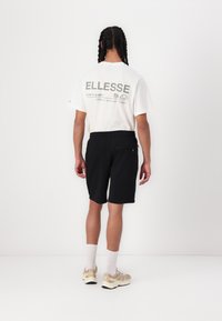 Svarte shorts og en hvit t-skjorte med grønn tekst og logo på ryggen. Modellen står med ryggen til kameraet, iført beige joggesko.