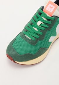 Green mesh and suede sneaker with a textured surface, featuring a pink logo tab and gum rubber sole. Laces are also green.