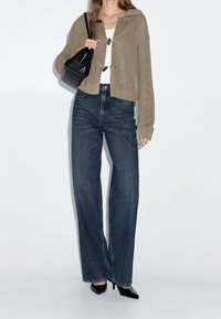 Cardigan tricoté beige court avec capuche, jean taille haute bleu foncé, talons noirs à bout pointu, et petit sac à main noir.