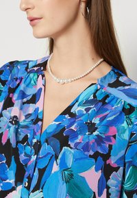 Blusa floreale blu con maniche a palloncino corte, sfondo nero e collana di perle. Presenta vivaci motivi di fiori blu, rosa e verdi.