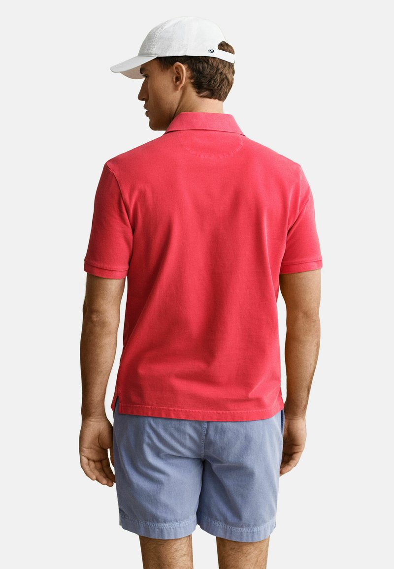 Mann mit weißer Kappe, rotem kurzärmligem Poloshirt und hellblauen Shorts, stehend mit dem Rücken zum Betrachter vor einfarbigem Hintergrund.