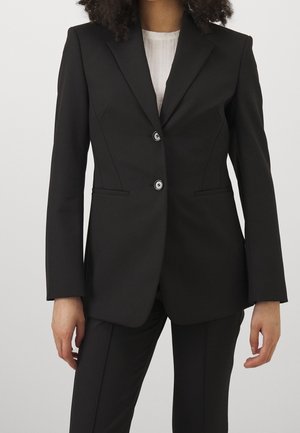 Vrouw die een nauwsluitend zwart colbert draagt met inkepingrevers, twee knopen aan de voorkant, bijpassende zwarte broek en een witte geribbelde top eronder.