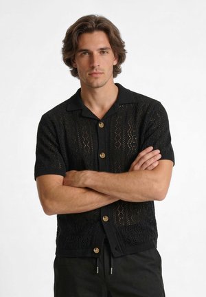 Noor mees keskmise pikkusega pruunide juustega, musta lühikeste varrukatega kootud nööbitava särgiga, käed ristatud, valge tausta ees seismas.