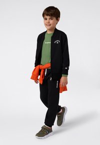 Niño sonriente caminando, vistiendo una chaqueta negra con cremallera, camiseta verde de Champion, pantalones de chándal negros, zapatillas color oliva y un sudadera naranja atada a la cintura.