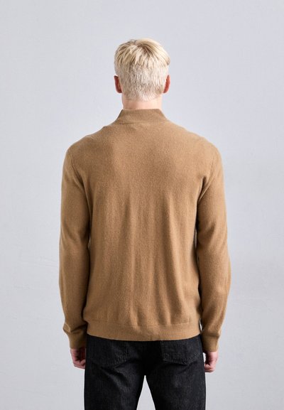 Blondiem matiem cilvēks ar muguru, tērpies kamieļa krāsas džemperī un melnos biksēs, stāvot vienkāršā gaiši pelēkā fonā.