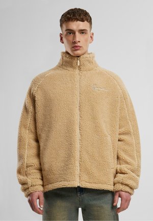 Vaaleanbeige fleece-takki, jossa on korkea kaulus, koko edestä avattava vetoketju ja ribbaantuneet hihansuut. Takissa on hienovaraista valkoista ommelta ja brodeerattu logo rinnassa.